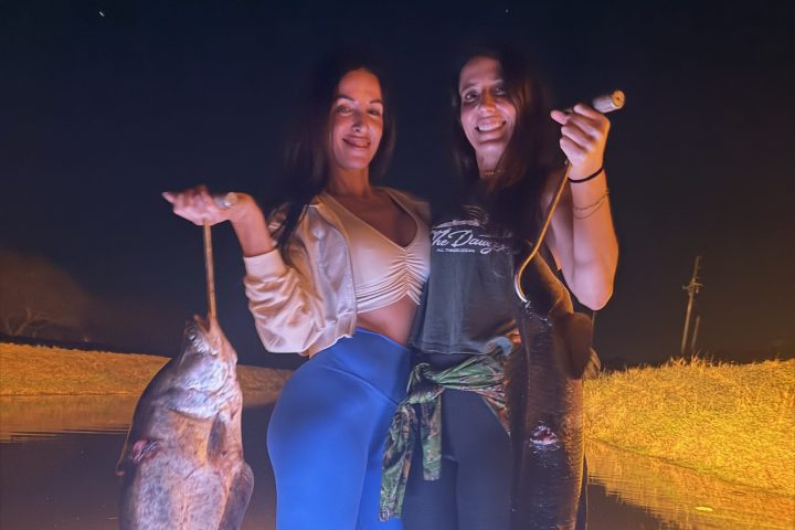 Two people on a boat holding large fish at night, illuminated by blue light.