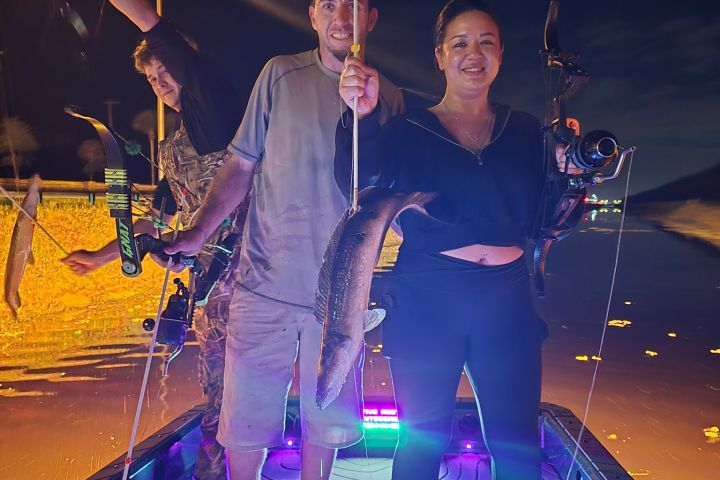 Three people on boat at night with bows and a caught fish.