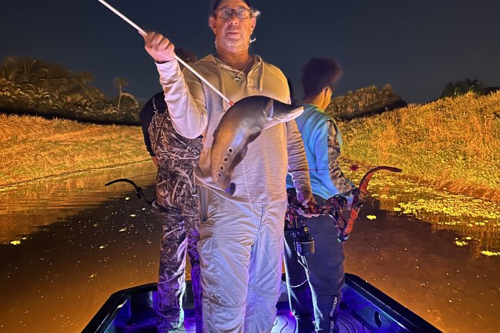 Three people on a boat at night, one holding a fish on a pole.