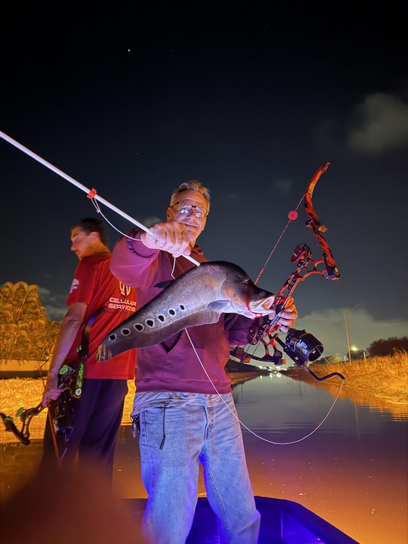 Two people on a boat at night, holding a large fish with a bowfishing setup.