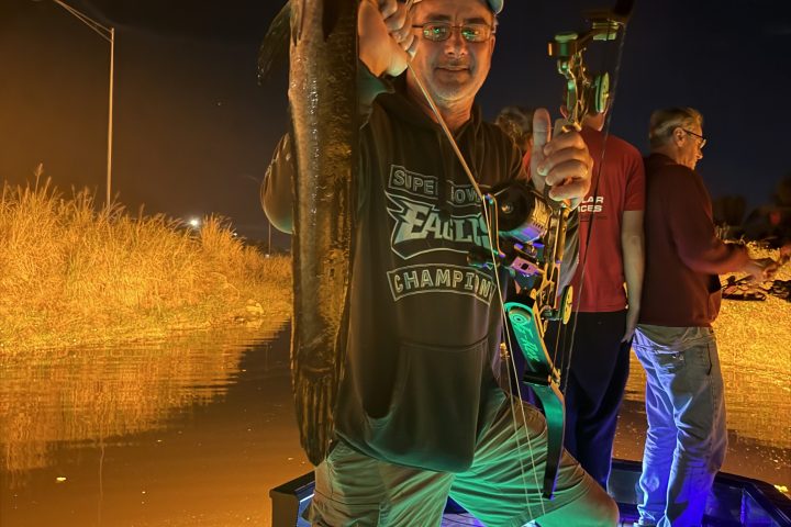 Man on a boat holding a fish and a bow at night with orange light in the background.