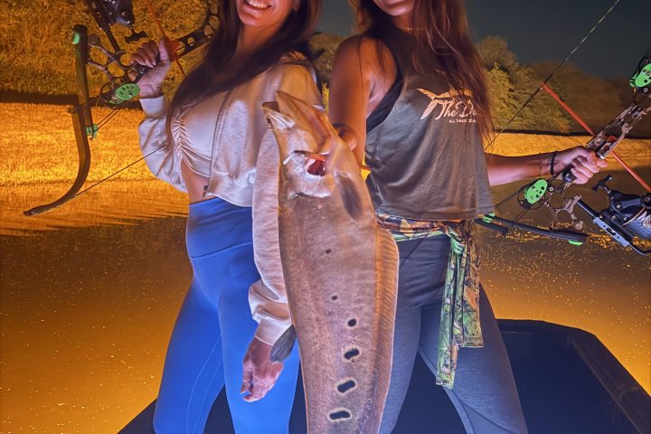 Two people smile holding a large fish at night, each with a bowfishing bow.